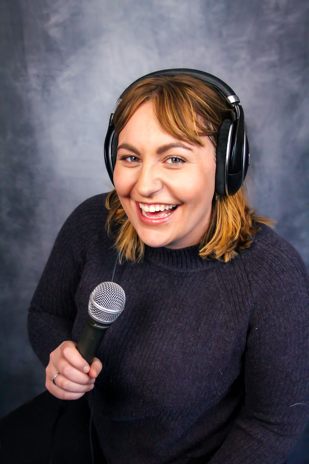Image of a smiling caucasian woman wearing over-ear headphones and a purple sweater in front of a grep backdrop. She has should-length brown hair and is holding a microphone.